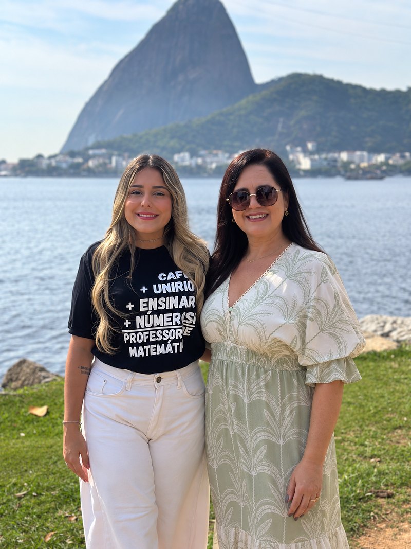Juliana e Alessandra, mãe e filha, lado a lado e sorrindo, com o mar e o Pão de Açúcar ao fundo