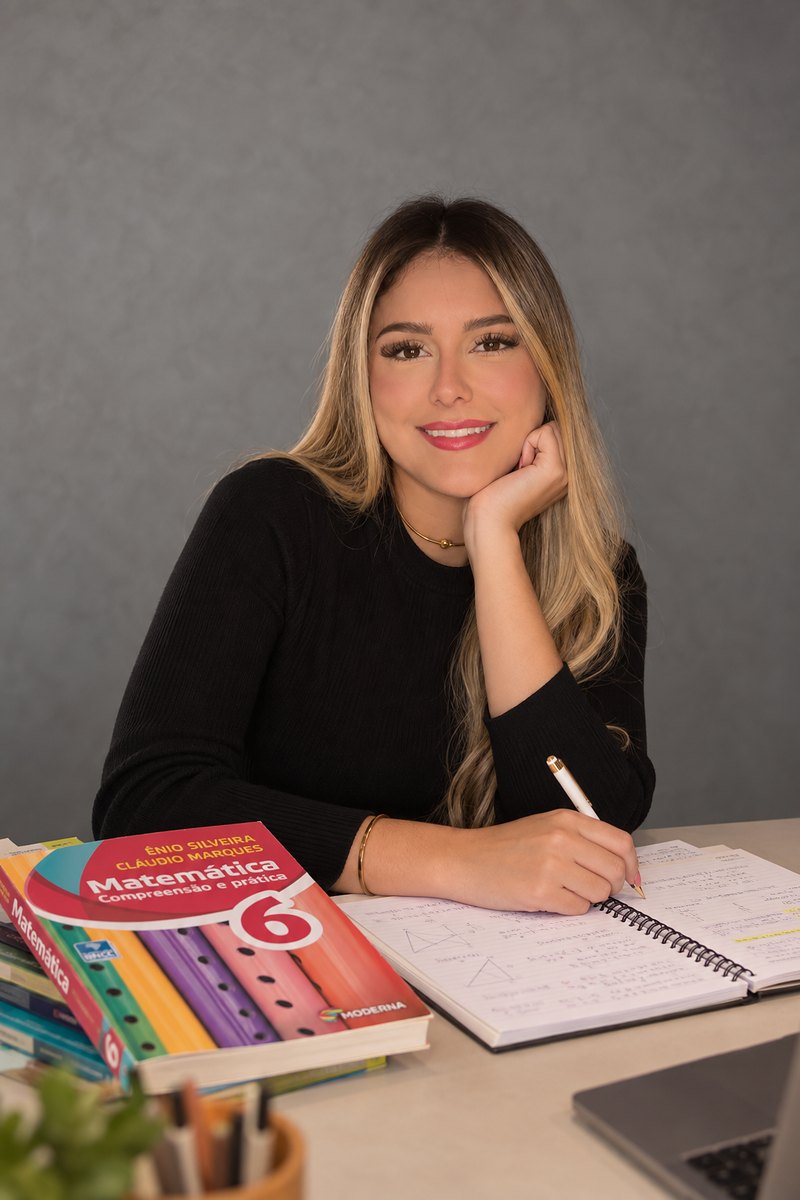 Juliana Oliveira, professora de Matemática, sorrindo à mesa de estudo, com livros e caderno